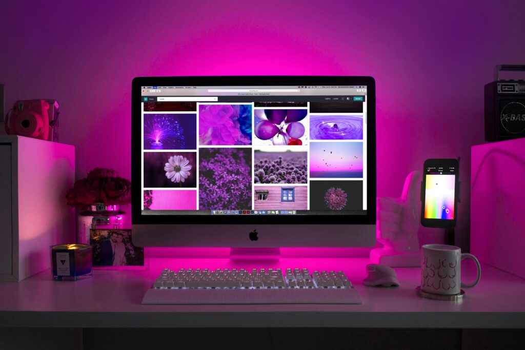 A computer monitor displays a grid of images on a desk with a keyboard and phone, illuminated by purple lighting.
